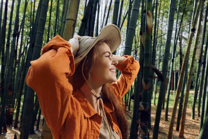 Woman in straw hat smiling in a bamboo forest, nature portrait of a happy traveler enjoying sunlight and greenery, outdoor relaxation and peaceful exploration among tall trees.
