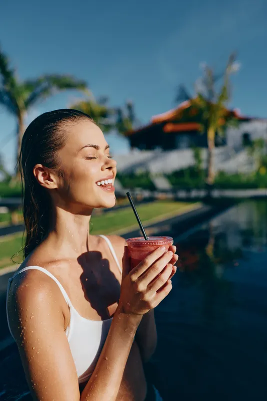 Relaxed woman by outdoor pool sipping a pink smoothie with straw, enjoying sunshine, tropical resort vibe, fresh breeze, casual swimwear, carefree mood, water reflection, holiday scene, peaceful