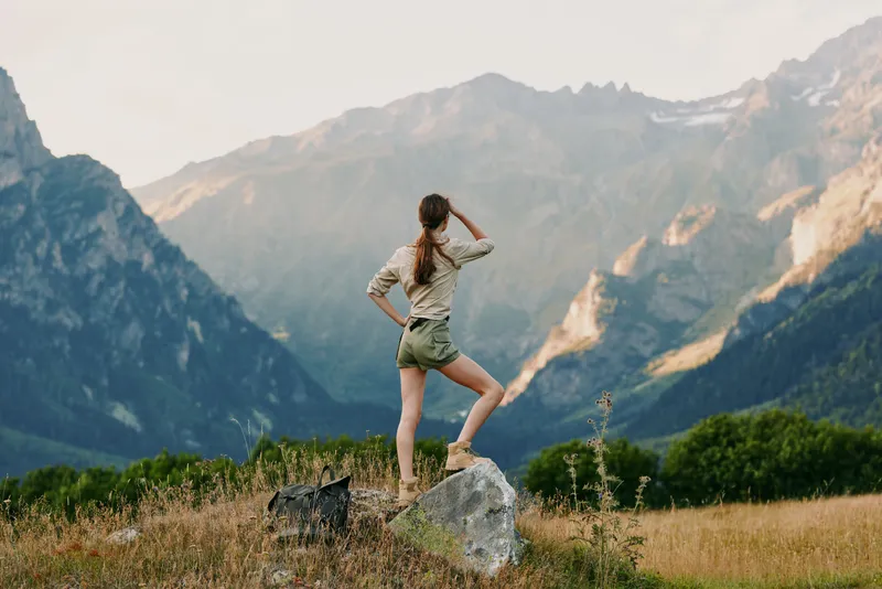 Young woman in casual clothes standing on a rock in a mountain field looking at distant peaks. Outdoor lifestyle, adventure, freedom, and nature concept.