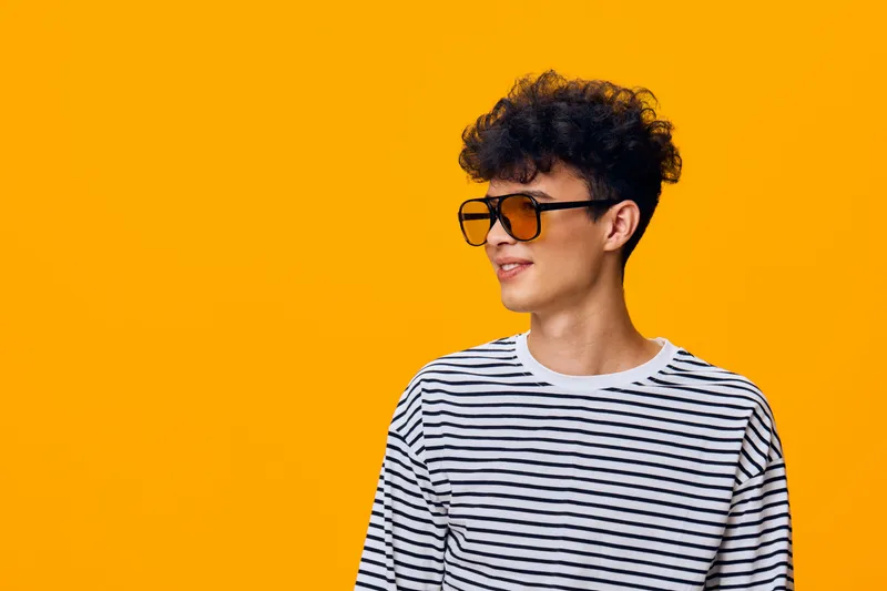 Young man wearing striped shirt and stylish sunglasses looking sideways with a cheerful expression against bright orange background, showcasing modern casual fashion.