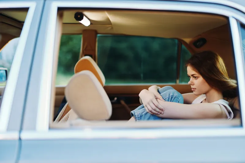 Sad young woman in casual outfit sitting in the backseat of a car, expressing deep emotions and reflection amidst a blurred green background.