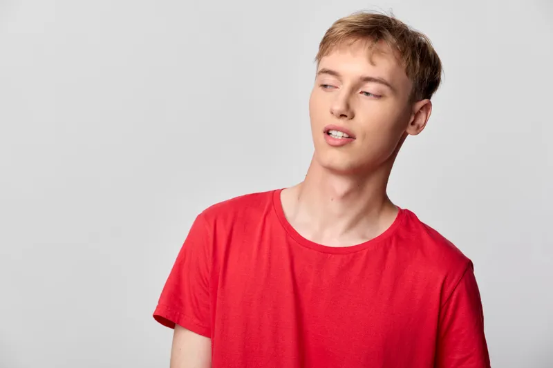 Young man in a red T shirt stands against a neutral backdrop with a relaxed pose and tilted head, conveying casual style, everyday charm, and a candid fashion moment