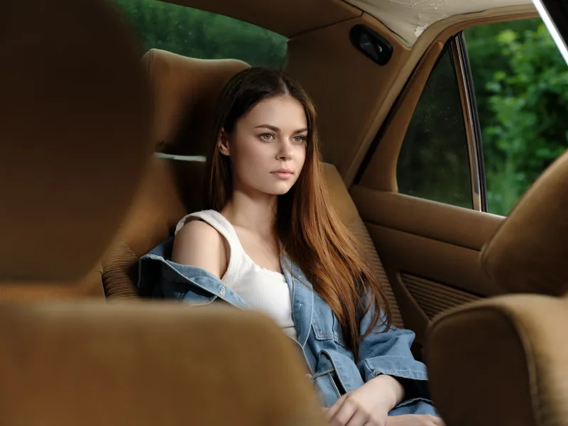 Young woman in a denim jacket sitting in a classic car with a contemplative expression, surrounded by a soft green background, conveying a sense of nostalgia and freedom.