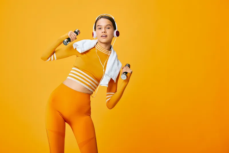 Woman fitness sport in a vibrant orange outfit, performing a dynamic workout with dumbbells, towel over shoulders and headphones against a bold orange backdrop; energetic exercise scene of athletic