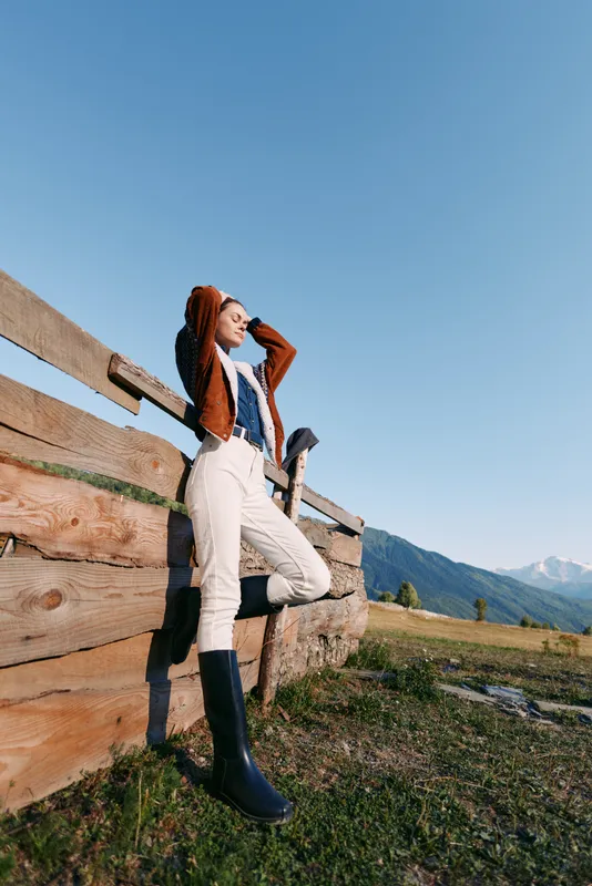Woman leaning on wooden fence in mountains meadow, wearing fashion boots and casual outfit, outdoor countryside portrait with clear sky and relaxed travel lifestyle.