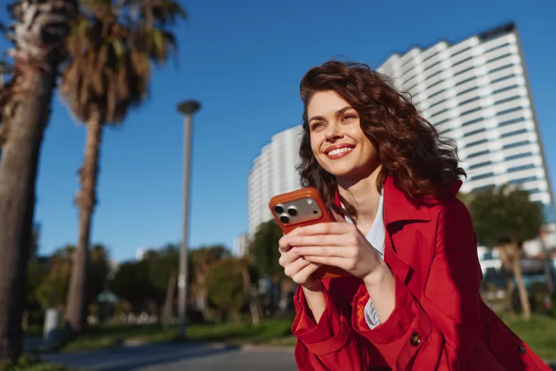 Young woman with curly hair wearing red jacket smiles holding smartphone outdoors with palm trees and modern building in background on sunny day.