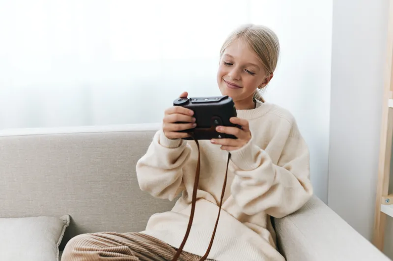 Authentic moment of a young student at home, playing a handheld game on a cozy couch, soft natural light, balanced lifestyle scene.