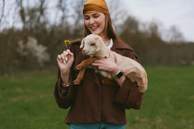 Joyful woman outdoors holding a baby goat, smiling as she offers a yellow flower. Casual jacket, headscarf, and denim meet a grassy field in soft natural light, capturing a warm lifestyle moment.