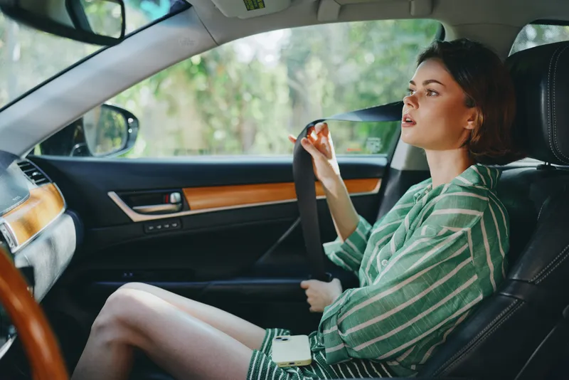 A woman driver sits in a modern car, gripping the steering wheel and gazing ahead as sunlight filters through the leafy interior.