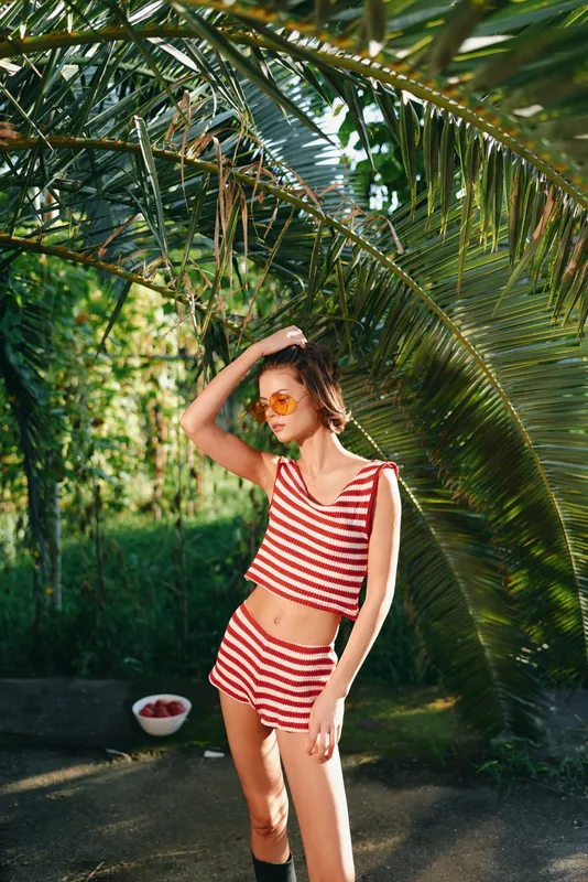 A stylish young woman stands outdoors in a sunny garden, wearing a red and white striped two piece swimsuit and sunglasses, posing confidently with a relaxed, vibrant vibe.