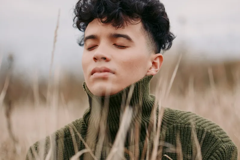 Close-up portrait of a young person with eyes closed in a windswept field, conveying authenticity and quiet realism through natural light, tall grass foreground, textured knitwear and serene,