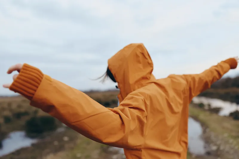 Woman outdoors lifestyle, hooded jacket and orange raincoat, arms stretched wide, wind in the hood, cloudy sky, travel moment, scenic nature landscape by water, carefree relaxed smile