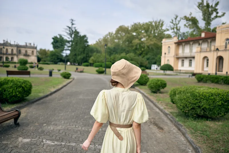 Woman in pale dress and wide hat walks along a gravel path in a manicured garden, with historic buildings visible in a serene estate setting