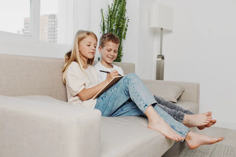 Two children sitting barefoot on sofa engaged in writing and drawing at home with natural light and modern interior decor.