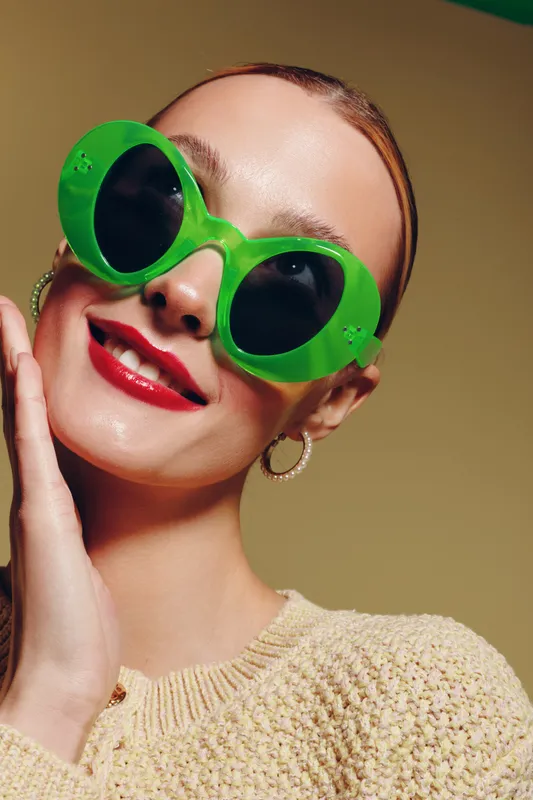 Close-up of a smiling woman wearing bright green sunglasses and red lipstick. She touches her face gently, wearing hoop earrings and a beige knitted sweater. Fashion and beauty concept.