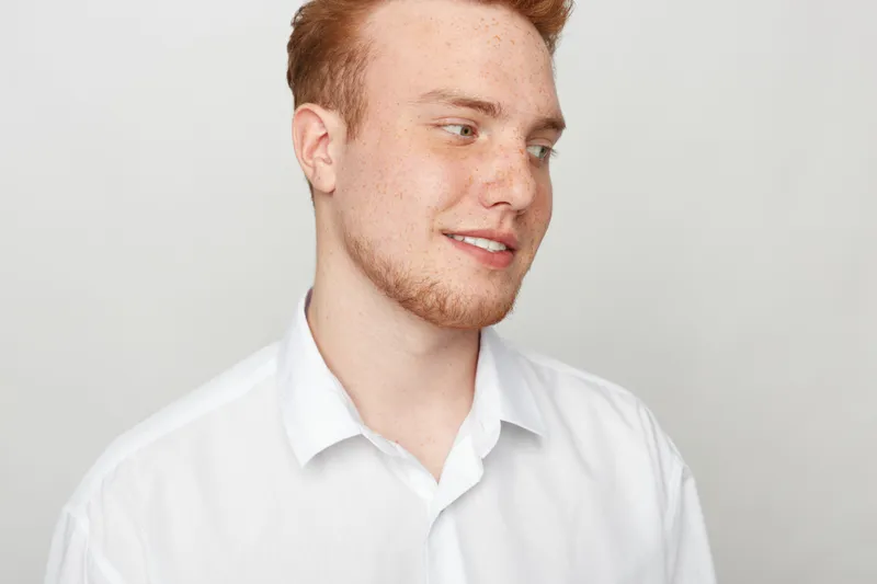 Young man with red hair and beard wearing white shirt looks sideways with slight smile. Studio portrait on light background. Concept of casual style, youth, and natural expression.