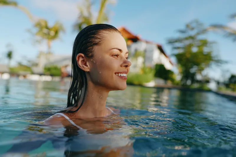 Woman with smile in a pool under tropical sunshine, capturing a relaxed lifestyle and wellness moment. Clear water reflections enhance leisure, vacation vibe, and self care in bright daylight.