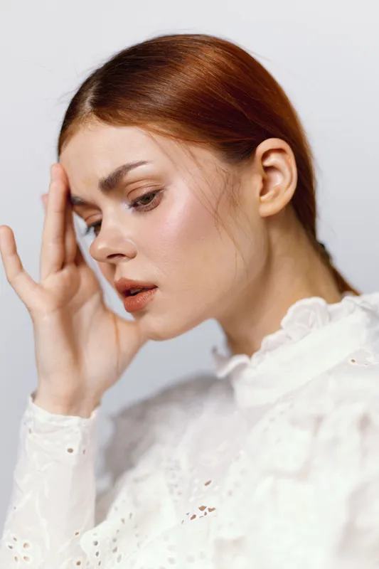 Young woman with red hair in a white blouse, expressing concern and contemplation against a soft, neutral background, capturing emotions of distress and reflection