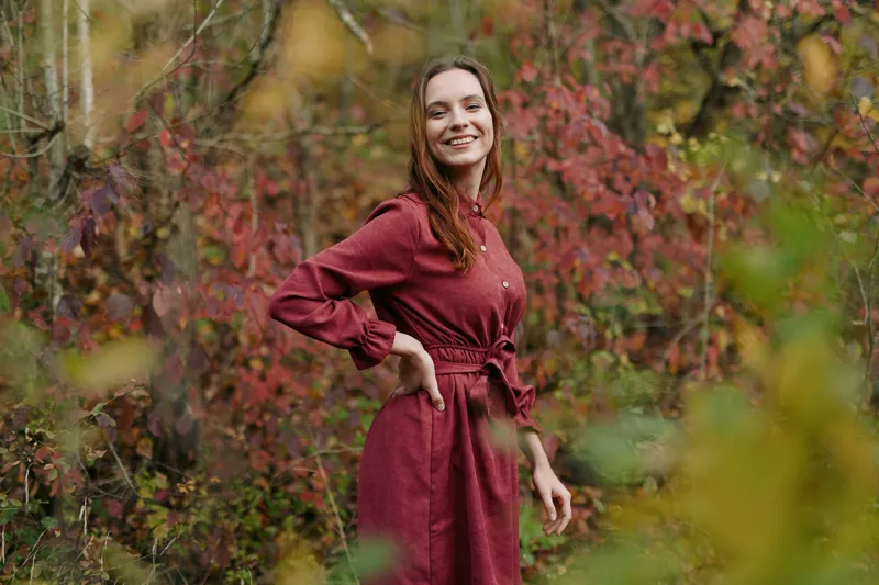 A smiling young woman in a simple red dress stands among autumn foliage, photographed with genuine emotion and natural composition to convey authenticity and accurate, believable expression.