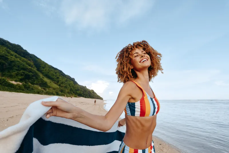 Young woman with curly hair smiles on a sunny beach, wearing a striped swimsuit and holding a towel, creating a carefree summer mood