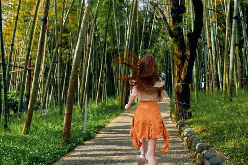 Woman running along a path in a bamboo forest, back view wearing an orange skirt, summer nature scene with green trees, motion blur and sunlight on a peaceful outdoor walkway.