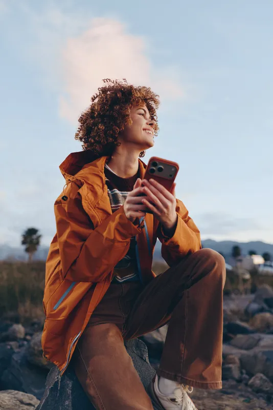 Woman lifestyle portrait outdoors on rocks, orange jacket, curly hair, smiling while holding phone, looking up at sky and clouds, casual summer daylight, gentle wind