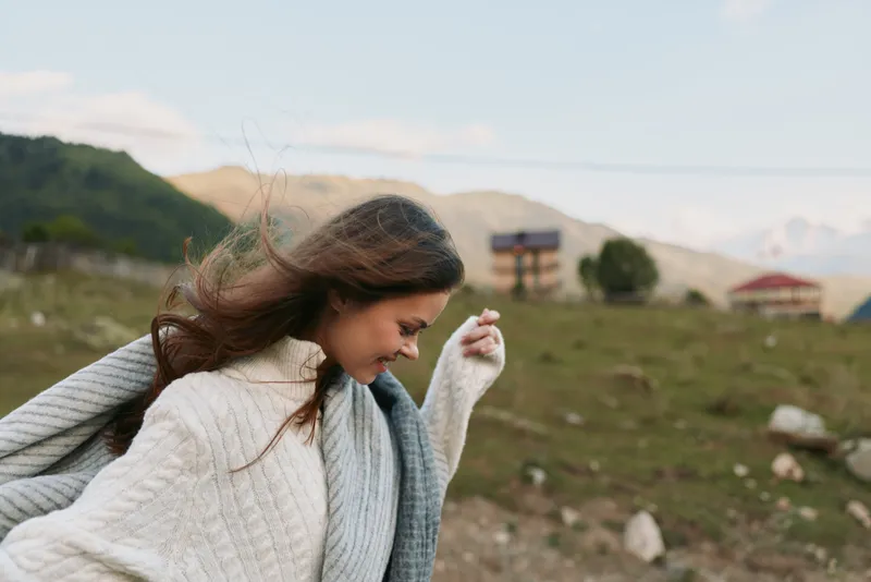 Casual outdoor portrait of a smiling woman in a cozy sweater, sunlit landscape with mountains, light breeze, candid lifestyle moment, natural colors, relaxed posture.