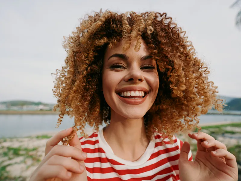 Joyful portrait of a young woman with curly hair smiling broadly outdoors by a lake, wearing a red and white striped shirt, exuding vibrant energy.
