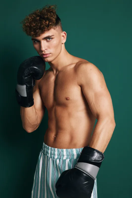 Young male boxer with curly hair wearing black gloves and striped shorts posing in fighting stance against dark green background in studio lighting.
