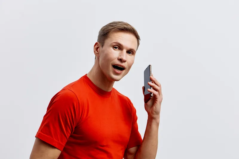 Smiling man wearing bright red T-shirt holding smartphone and looking into camera, casual pose, friendly facial expression, isolated on plain white background, studio portrait. People lifestyle concep