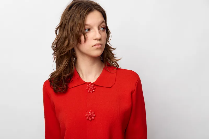 Young woman with curly brown hair wearing a red knitted sweater looks to the side with a neutral expression. Studio portrait on white background with soft lighting and casual style.