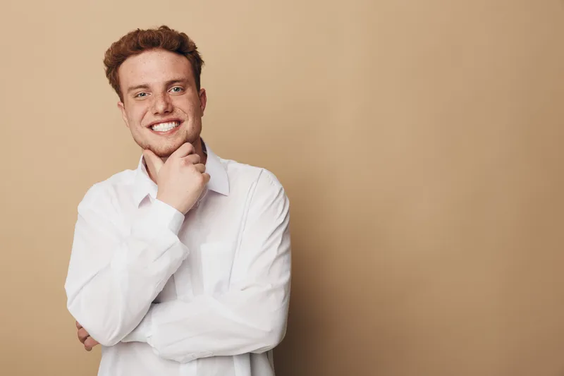 Smiling young man with red hair wearing white shirt poses with hand on chin against beige background. Concept of confidence, casual style, and positive emotions in studio portrait.