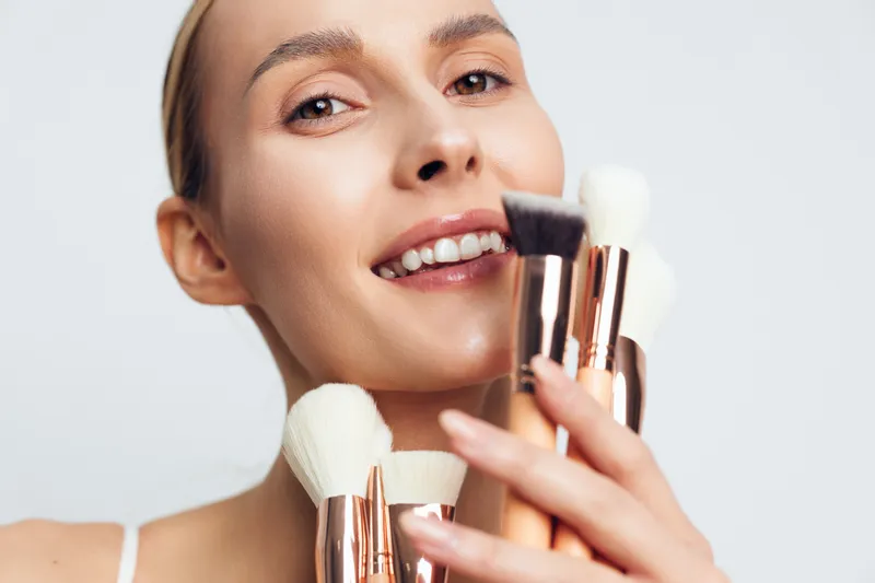 A smiling young woman holds a set of makeup brushes close to her face, showcasing a fresh beauty routine in a clean studio portrait with soft lighting.