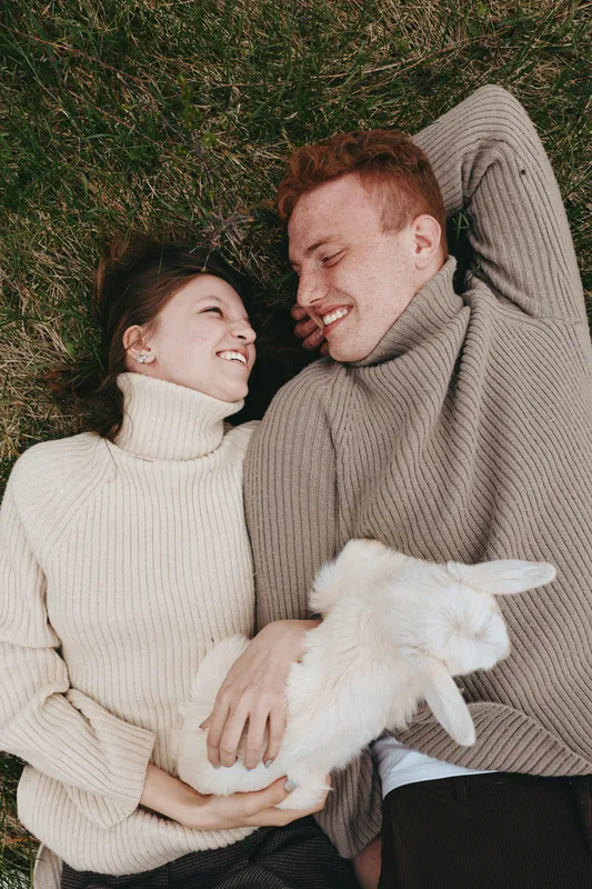 Smiling couple lying on grass in cozy turtlenecks, holding a fluffy goat kid between them. Warm natural light conveys relaxed lifestyle, gentle affection, and countryside charm.