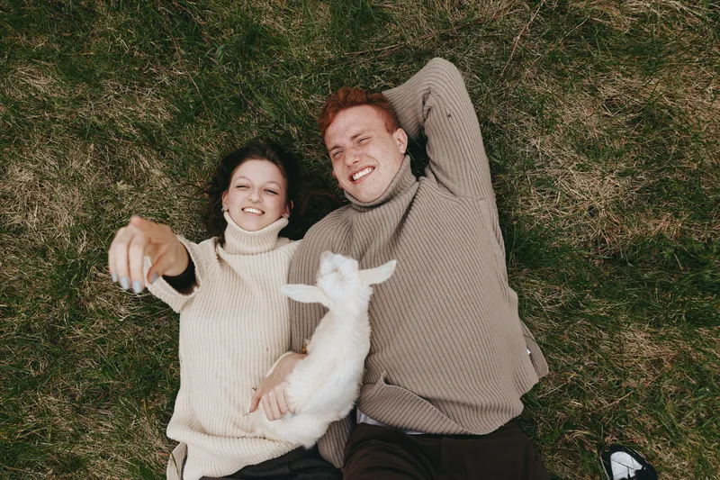 Playful couple outdoors lying on grass, smiling at camera with a white goat kid, warm knit sweaters, natural light, relaxed mood, rustic setting, candid moment, gentle breeze, vibrant greens,