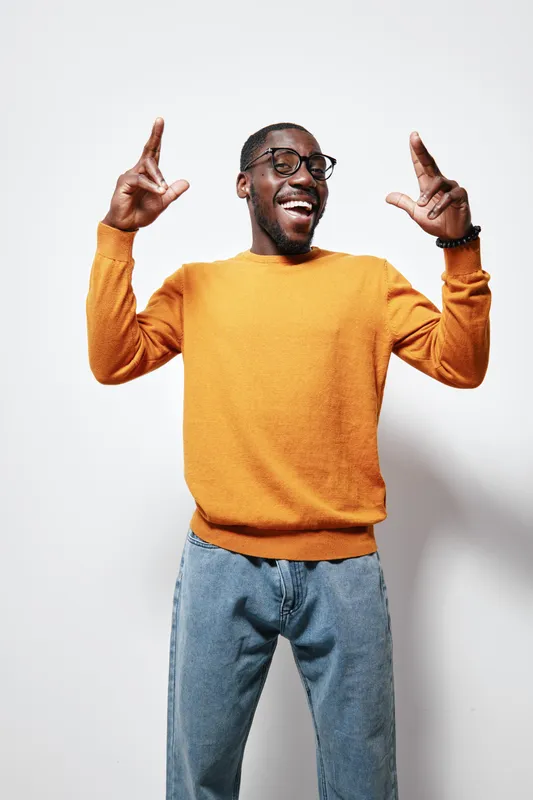 African American man with dark skin wearing glasses and orange sweater smiling and posing with hands up against white background in casual style.