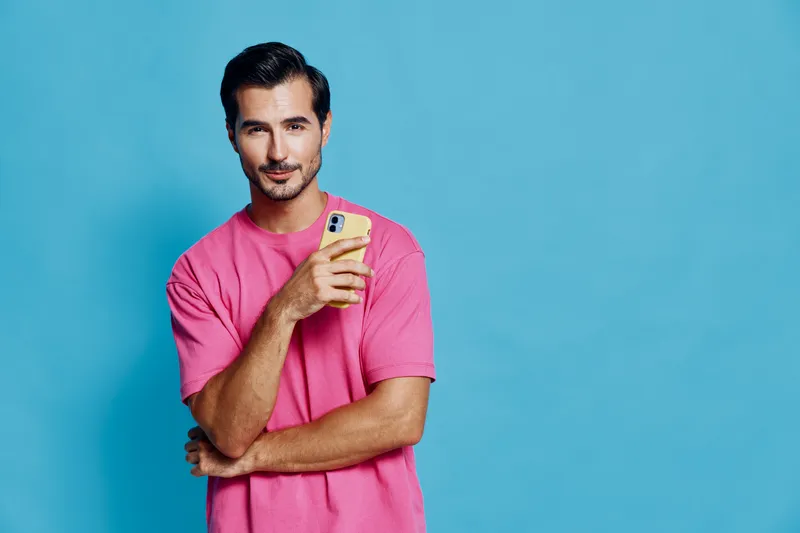 Confident smiling man in pink t-shirt holds smartphone while posing against vibrant turquoise background, exuding modern lifestyle and tech-savvy vibe.