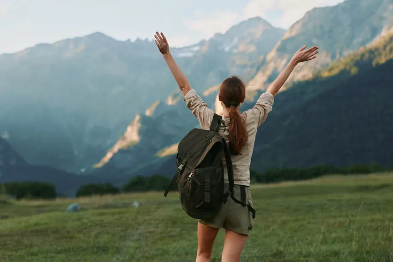 Woman hiker with backpack raising hands enjoying mountain landscape outdoors, casual clothing, summer day, green grass field, clear sky, active lifestyle concept