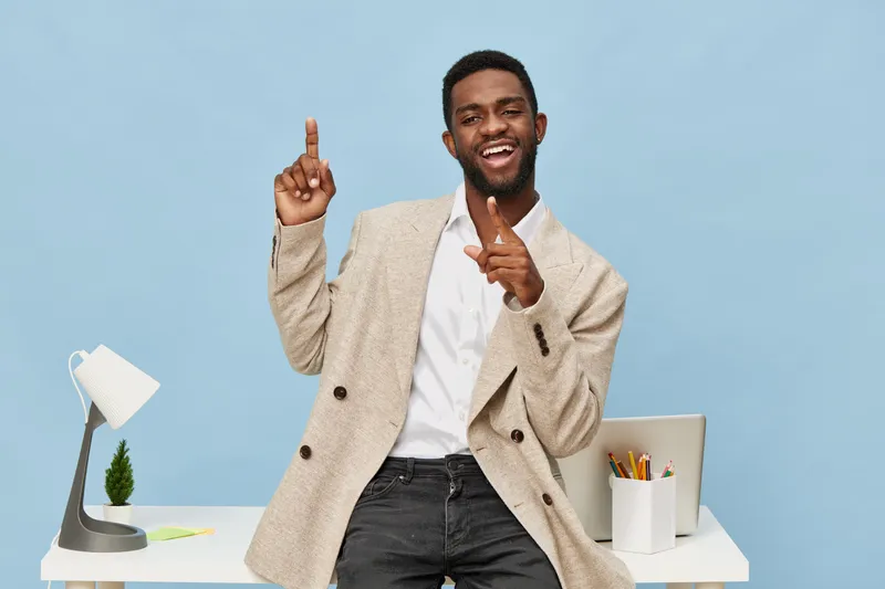Confident young man with beard smiling and pointing upward while standing in office with blue background, desk lamp, plant, and stationery, casual professional style. People lifestyle concept