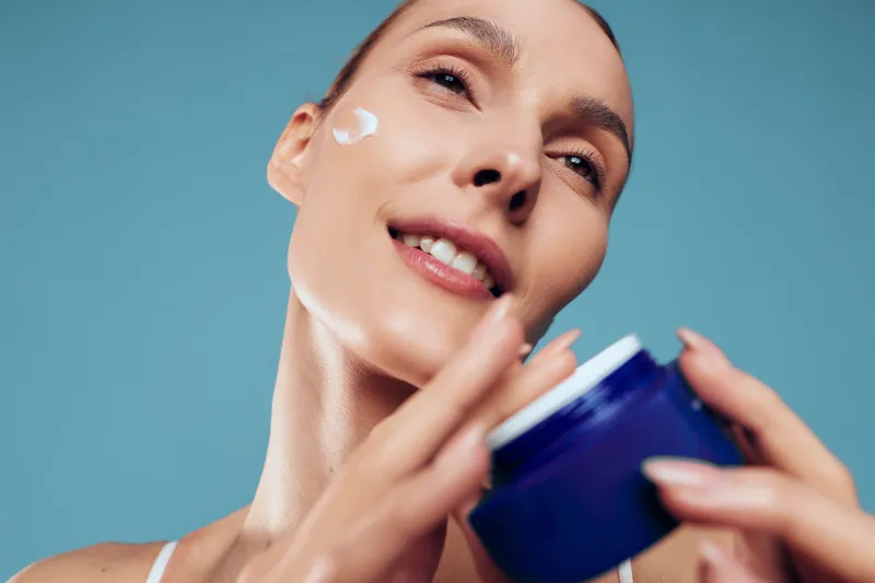 Young woman with short hair applies moisturizer on her cheek while holding a blue jar against a plain blue background, showing skincare and self-care routine with a gentle smile.