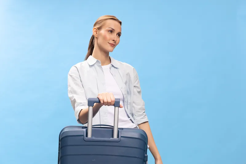 Young woman in casual clothes holding a blue suitcase handle, standing against a light blue background. Concept of travel, vacation, and summer break with luggage and tourism.