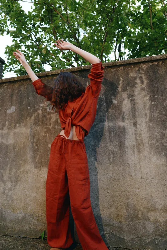 A woman in a burnt orange jumpsuit stands with arms raised, back to the camera, beside a weathered wall in an outdoor setting. A bold, energetic lifestyle moment