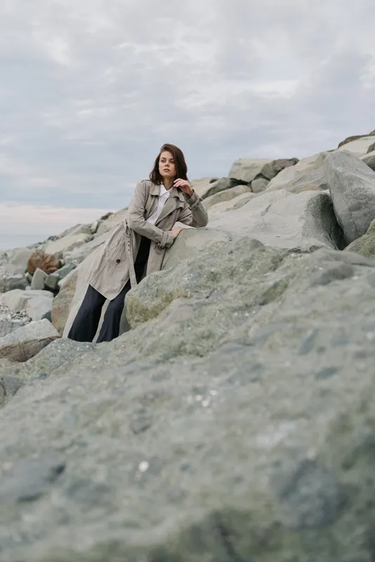 Young woman in trench coat stands on rocky shore under cloudy sky, posing thoughtfully. Concept of fashion, nature, and autumn mood with casual style and outdoor scenery.