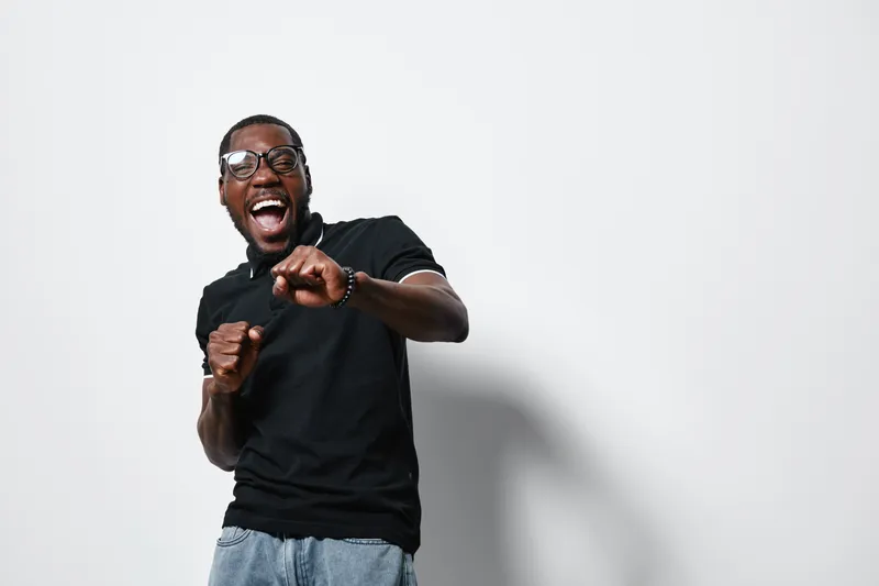 African American man with dark skin wearing glasses and black t shirt laughing and dancing against white background in casual style with joyful expression.