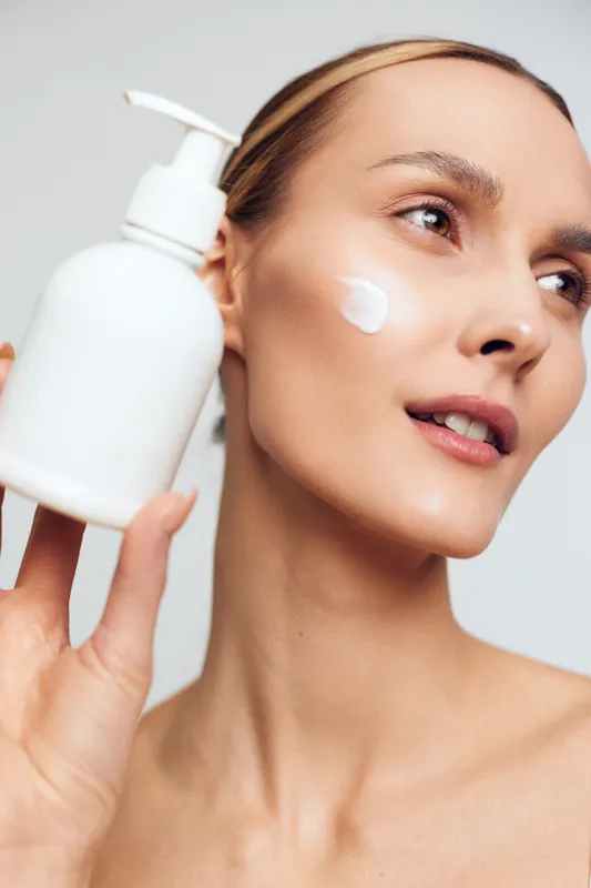 Smiling woman with smooth skin applying white face cream from a pump bottle. Closeup on a plain background. Concept of skincare, wellness, and selfcare. Soft lighting creates a warm mood.