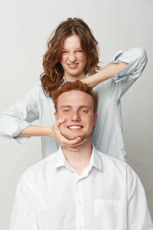 Playful young woman with curly hair teasing smiling redhead man in white shirts against plain background. Concept of friendship, fun, and casual interaction between friends.