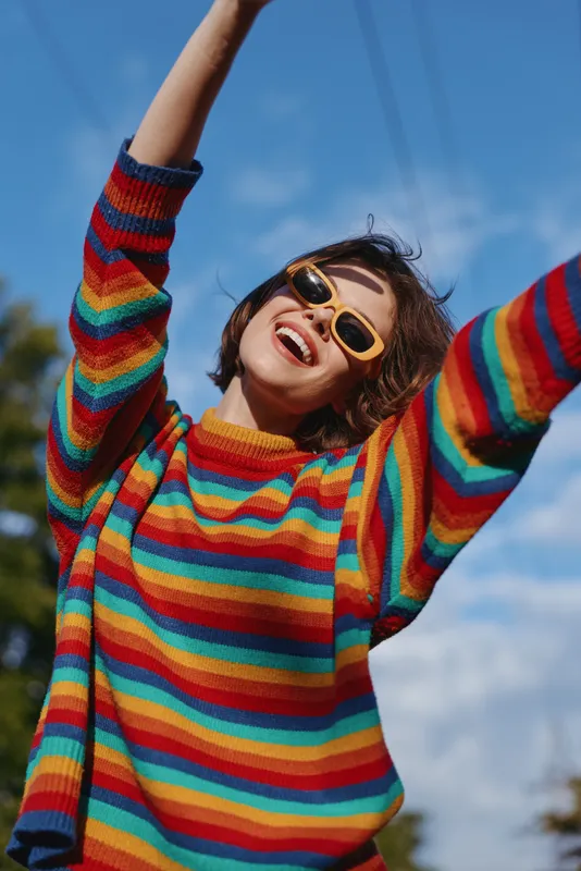 young woman in her 20s wearing a rainbow sweater and yellow sunglasses, smiling with arms up outdoors under blue sky; joyful casual outfit, short hair, lifestyle and fashion portrait.