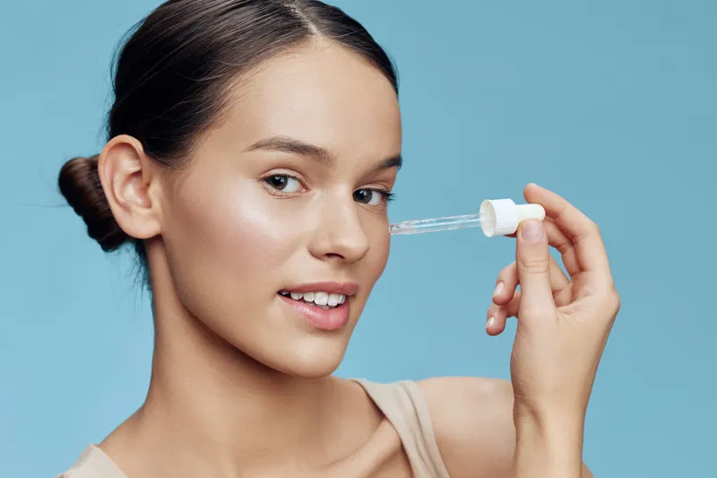 Young woman applying a serum to her face, showcasing healthy skin and beauty in a light blue background, promoting skincare and self care routines