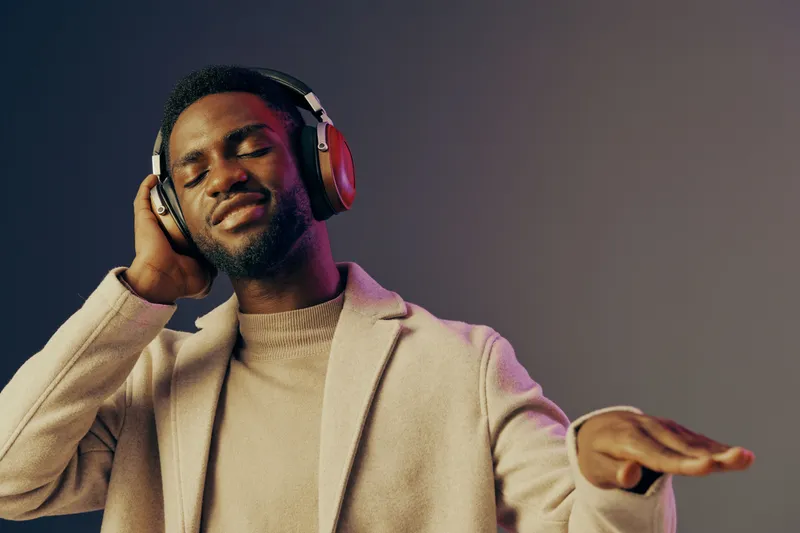 Young man enjoying music with headphones, wearing a stylish beige coat against a gradient background, showcasing a joyful and relaxed emotion
