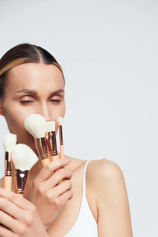 A young woman with closed eyes holds several white makeup brushes with rose gold handles near her face against a plain light background, concept for beauty and skincare.